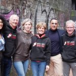 Let's Feed Brum Trustees stand side-by-side on a red brick canal path in Birmingham. In the background is a neutral brick wall covered in street art.